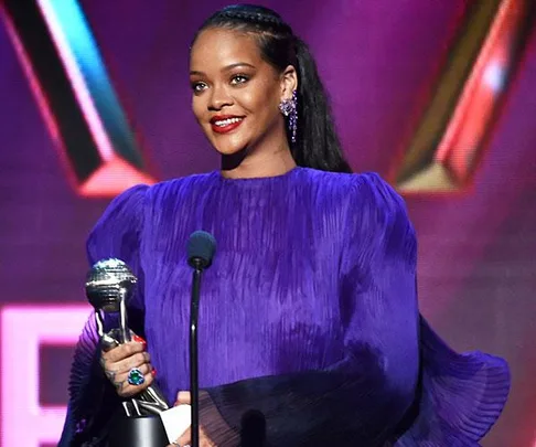 Singer in a purple dress smiles while holding a silver trophy on stage against a colorful backdrop.