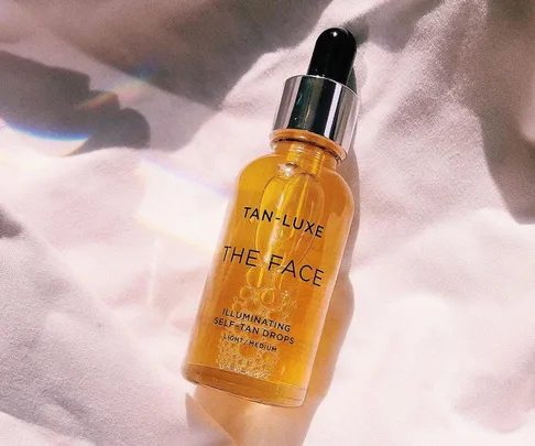 A bottle of Tan-Luxe The Face Illuminating Self-Tan Drops on a white fabric background.