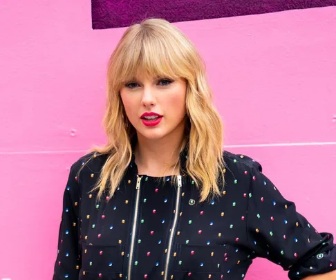 A woman with blond hair in a polka dot outfit stands against a pink wall.