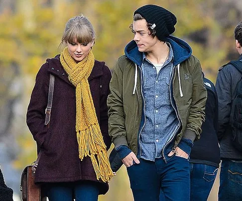 A woman in a yellow scarf and a man in a beanie walk together, talking, during a chilly day with autumn leaves in the background.