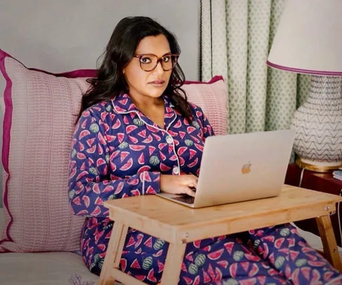 A woman in watermelon pajamas works on a laptop at home, sitting on a bed with a tray table.