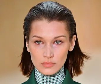 A person with slicked-back dark hair, wearing a green and black checkered outfit, looking directly at the camera.