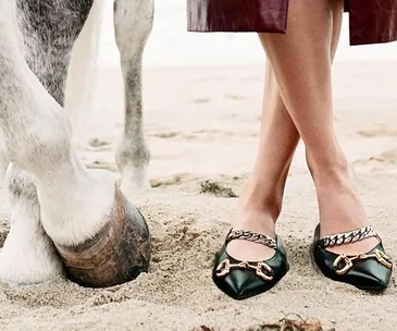 A woman in ballet flats stands on sand beside a horse's hoof, wearing a red skirt.