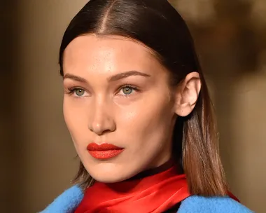 A model with straight brown hair, wearing red lipstick, a red scarf, and a blue outfit, poses on a runway.