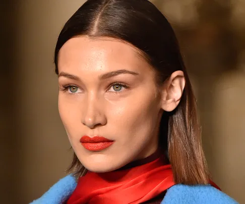 A model with straight brown hair, wearing red lipstick, a red scarf, and a blue outfit, poses on a runway.