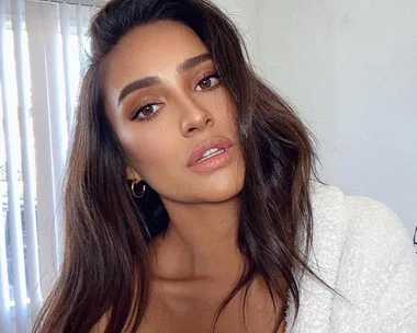 Woman with long dark hair and natural makeup, wearing a white textured top, poses in front of vertical blinds.