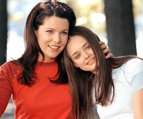 Two women, smiling and embracing, in casual attire with trees blurred in the background.