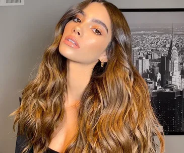 Woman with wavy brown hair posing in front of a black-and-white cityscape photo, wearing earrings and a black top.