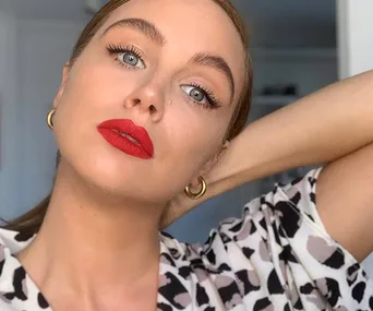 Woman with red lipstick, light eyes, and hoop earrings posing indoors, wearing a patterned top.