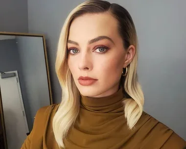 A person with blonde hair and a side part, wearing a brown turtleneck, poses in front of a mirror.