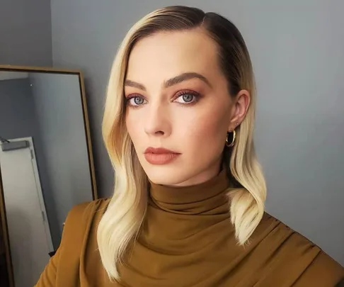 A person with blonde hair and a side part, wearing a brown turtleneck, poses in front of a mirror.