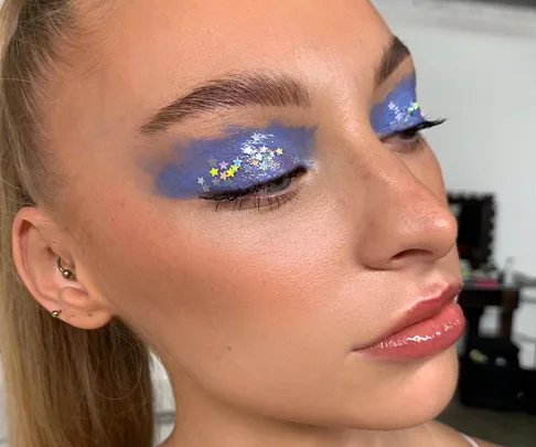 Woman with blue eyeshadow, decorated with tiny star shapes, and glossy lips, in a close-up shot.