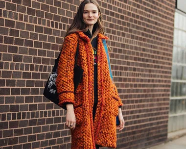 A woman poses in front of a brick wall wearing an orange textured coat and carrying a black tote bag, smiling.