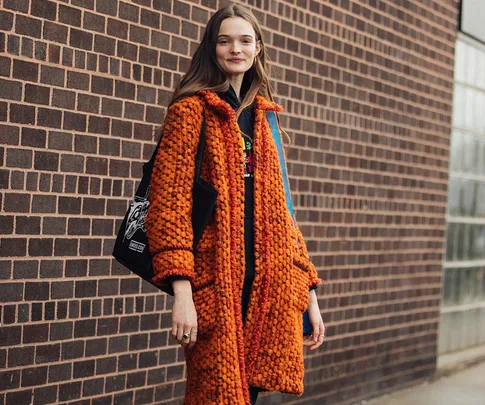 A woman poses in front of a brick wall wearing an orange textured coat and carrying a black tote bag, smiling.