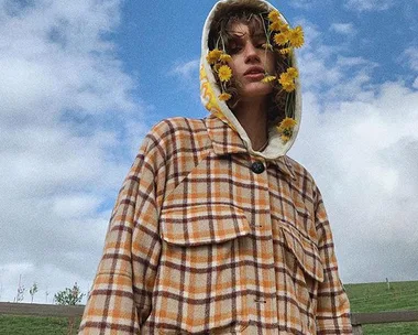 A person in a plaid coat with flowers around their face stands outdoors against a cloudy sky.