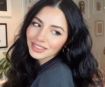 Woman with long, wavy, dark hair smiling with soft makeup, wearing a black top and gold hoop earrings.