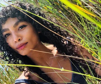 A woman with curly hair and a heart necklace amidst tall grass.