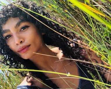 A woman with curly hair and a heart necklace amidst tall grass.