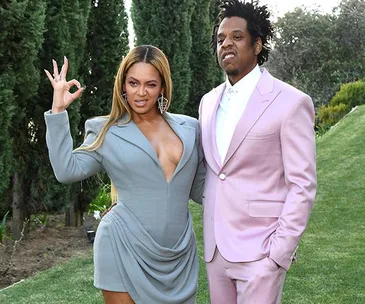 A couple poses elegantly outdoors; she wears a draped gray dress, and he dons a pastel pink suit, both expressing confidence.