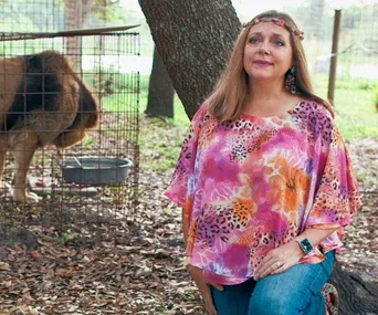 A woman in a colorful blouse kneels beside a lion in a cage outdoors.