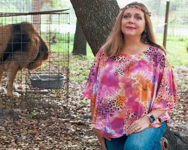 A woman in a colorful blouse kneels beside a lion in a cage outdoors.