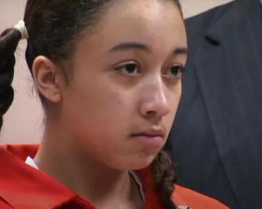 Young woman in an orange jumpsuit looks serious in a courtroom setting.