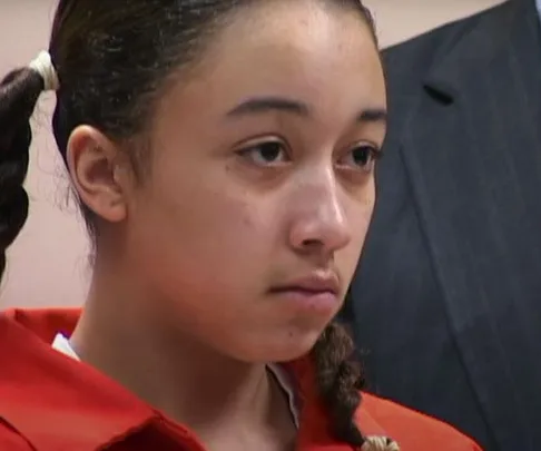 Young woman in an orange jumpsuit looks serious in a courtroom setting.