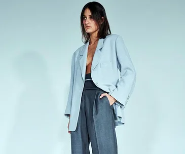 A model wearing a light blue oversized blazer with grey high-waisted trousers against a plain background.