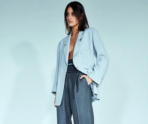 A model wearing a light blue oversized blazer with grey high-waisted trousers against a plain background.