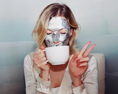 Woman with silver face mask holding a mug and making a peace sign.