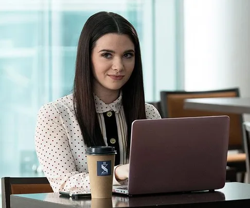 A woman sits at a table with a laptop and a coffee cup, wearing a white polka-dot blouse, smiling slightly.