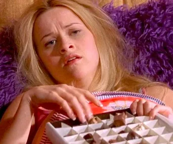 A person lying on a purple couch, looking upset while holding an open box of chocolates.