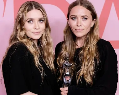 Two women with long wavy hair in black outfits, one holding a silver trophy, stand against a red and pink background.