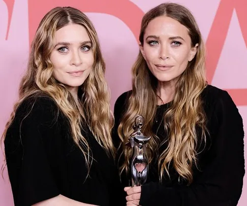 Two women with long wavy hair in black outfits, one holding a silver trophy, stand against a red and pink background.