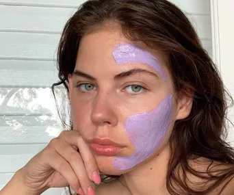 Woman with purple face mask, partially applied to forehead and cheek, resting her chin on her hand.