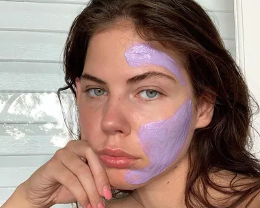 Woman with purple face mask, partially applied to forehead and cheek, resting her chin on her hand.