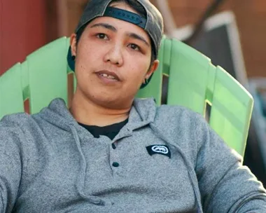 Person in a gray hoodie and backward cap sitting outside on a green chair.