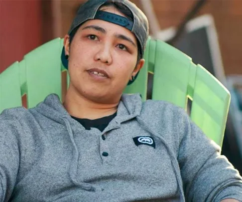 Person in a gray hoodie and backward cap sitting outside on a green chair.