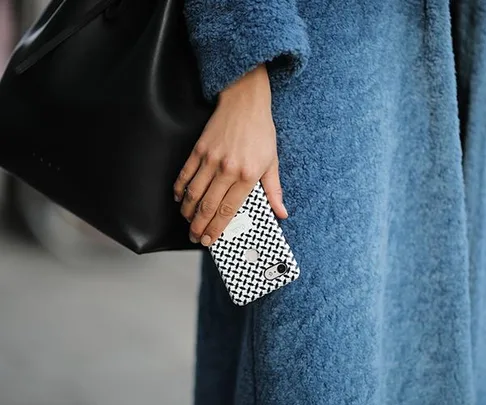 A person in a blue coat holding a patterned smartphone and a black bag.