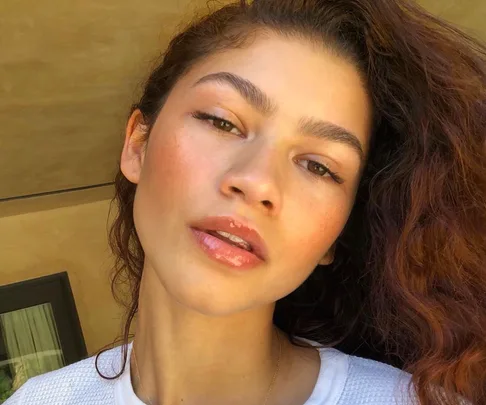 Close-up of a woman with curly hair, natural makeup, and a white sweater, looking directly at the camera.