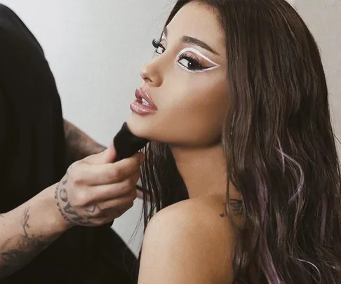 A person with styled long hair, bold white eyeliner, and glossy lips receives makeup touch-up.