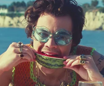 Person with curly hair, wearing large sunglasses, bites a watermelon slice by the ocean.