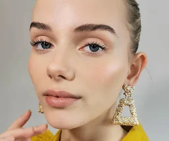 Close-up of a person wearing large gold, triangular earrings and a yellow top against a neutral background.