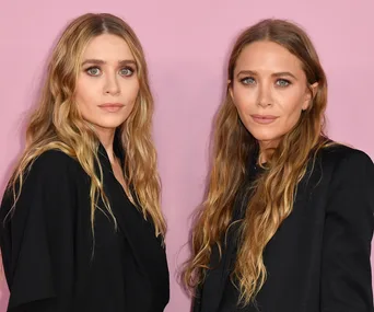 Two women with wavy, blonde hair stand against a pink background, both wearing black outfits.