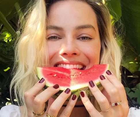 Woman smiling and holding a slice of watermelon in front of her face, with green leaves in the background.