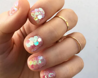 Close-up of a hand with iridescent hexagonal nail art and gold rings on fingers.