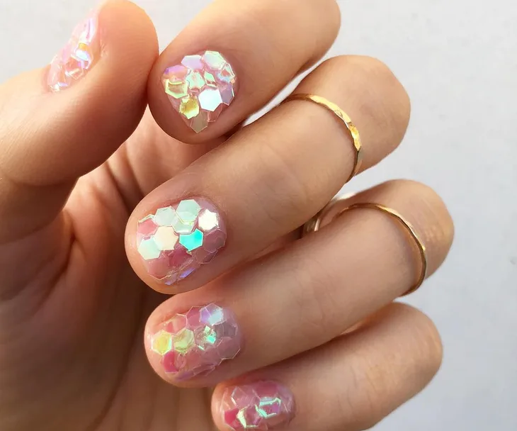 Close-up of a hand with iridescent hexagonal nail art and gold rings on fingers.