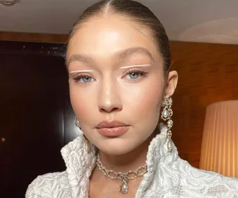 Woman in white outfit with pearl earrings and a necklace, makeup highlighting eyes, posed indoors.