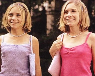 Young twins smiling outdoors, wearing pink tops, with matching shoulder bags and necklaces.