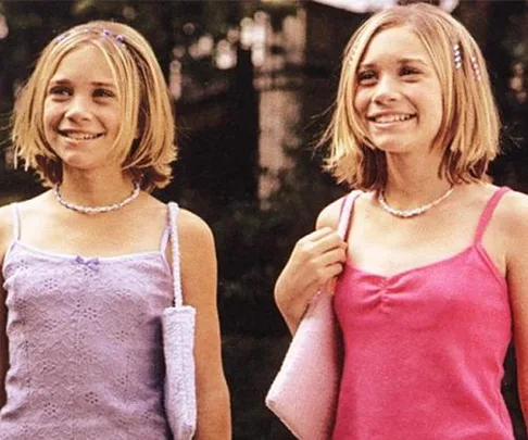 Young twins smiling outdoors, wearing pink tops, with matching shoulder bags and necklaces.